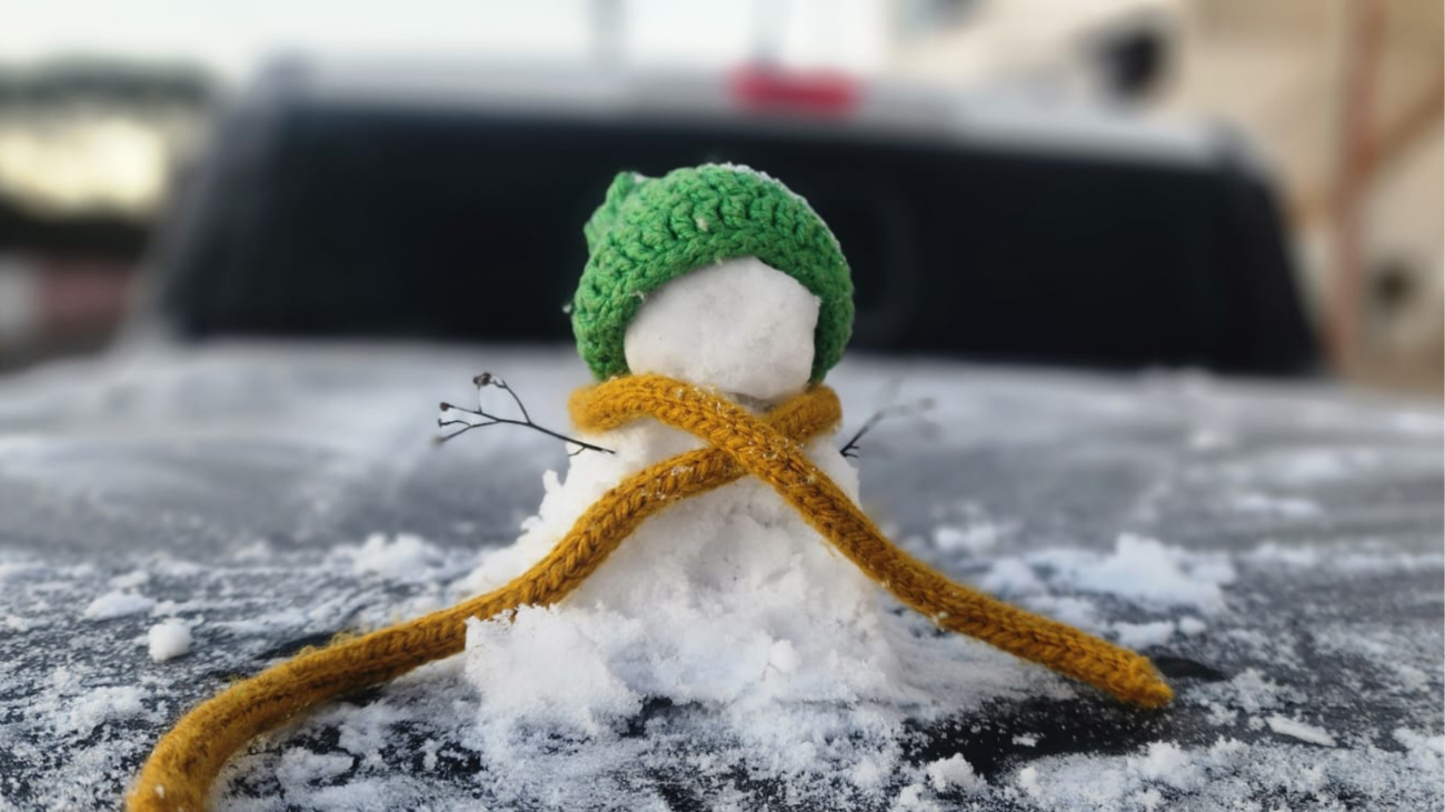 Pequeno boneco de neve com gorro verde e cachecol amarelo sobre carro coberto por neve durante inverno rigoroso